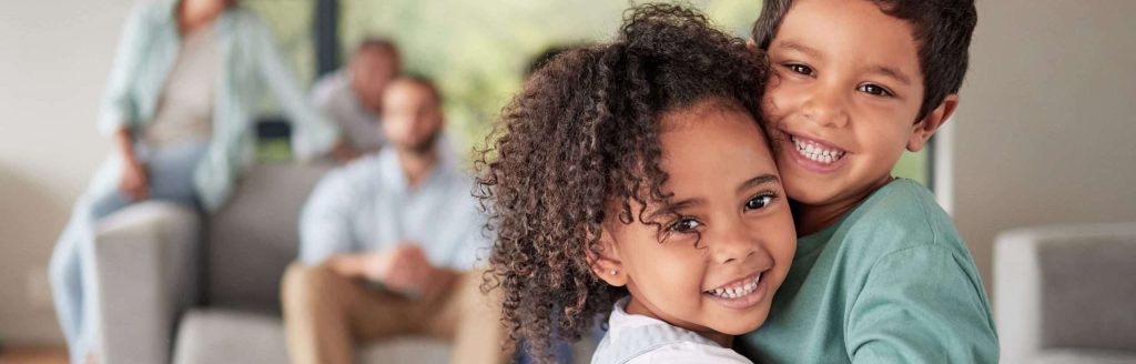 Girl and boy siblings hugging with blurred parents in the background.